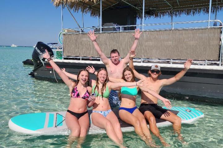 Five people smiling and posing on paddleboards in clear water near a boat under a blue sky.