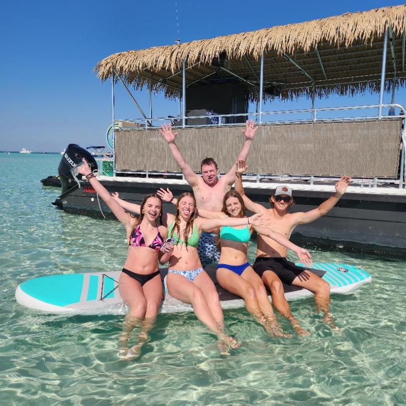 Five people smiling and posing on paddleboards in clear water near a boat under a blue sky.