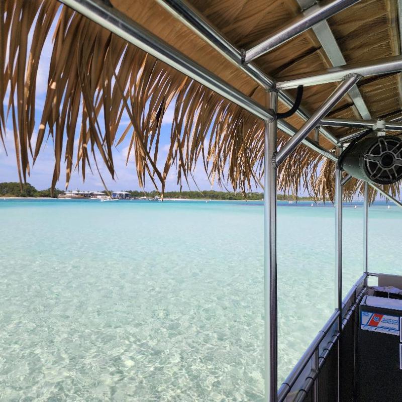 View from a palm-thatched boat of clear turquoise water and distant shore on a sunny day.