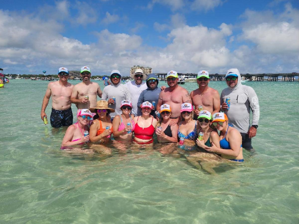 Group of people in swimsuits and colorful hats standing in shallow water holding drinks.