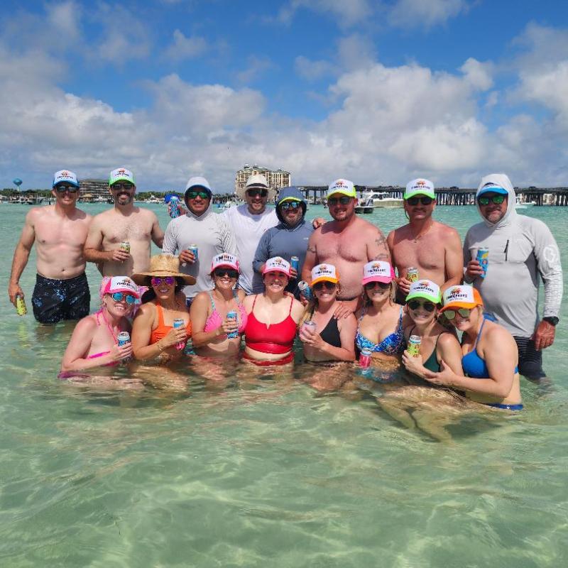 Group of people in swimsuits and colorful hats standing in shallow water holding drinks.