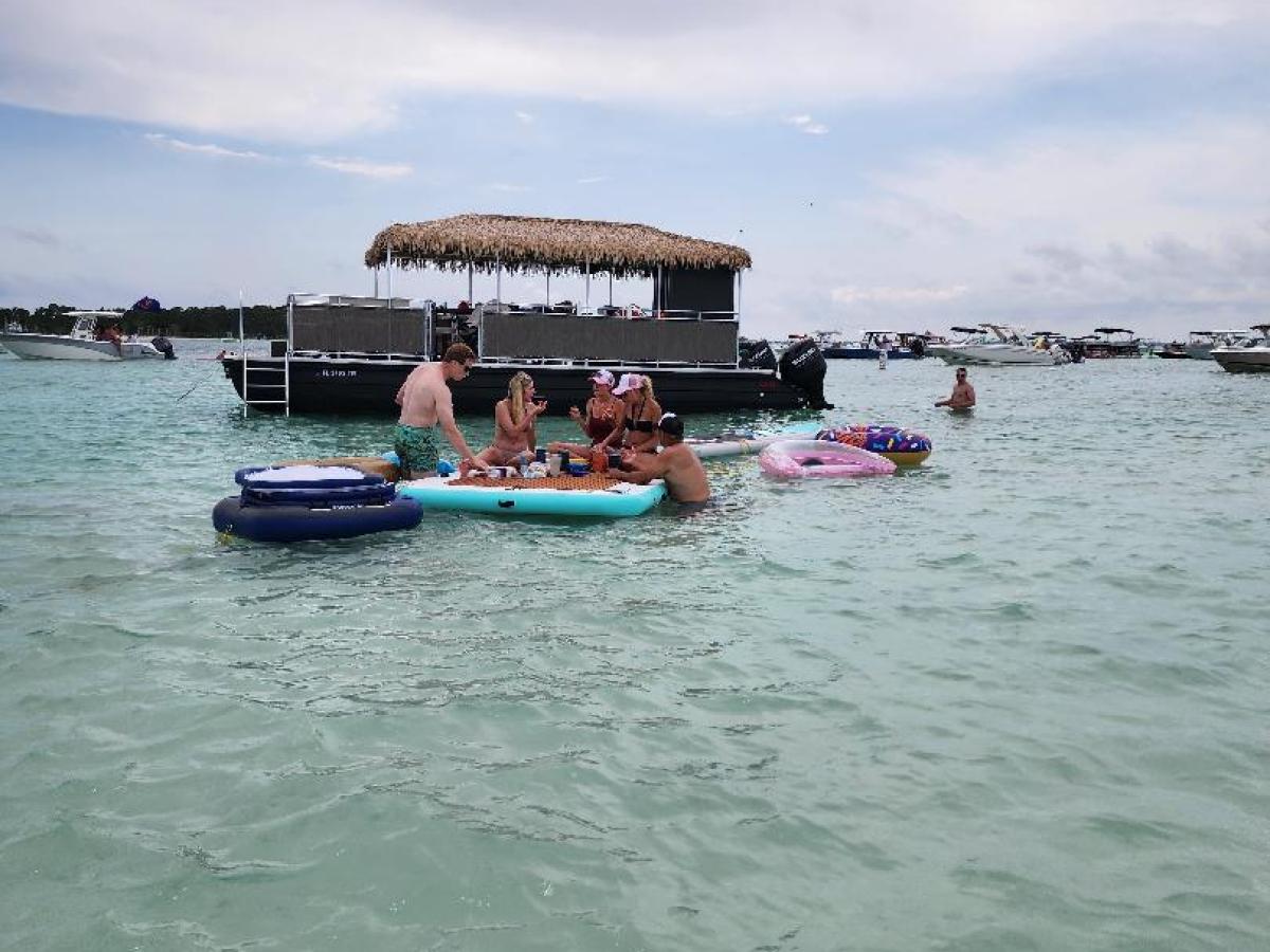 People relaxing on floating devices in clear water near a boat with a thatched roof.