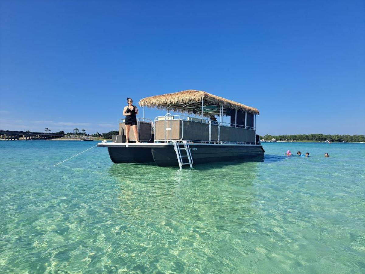 Person on tiki-style pontoon boat in clear water, swimmers nearby, under a clear blue sky.