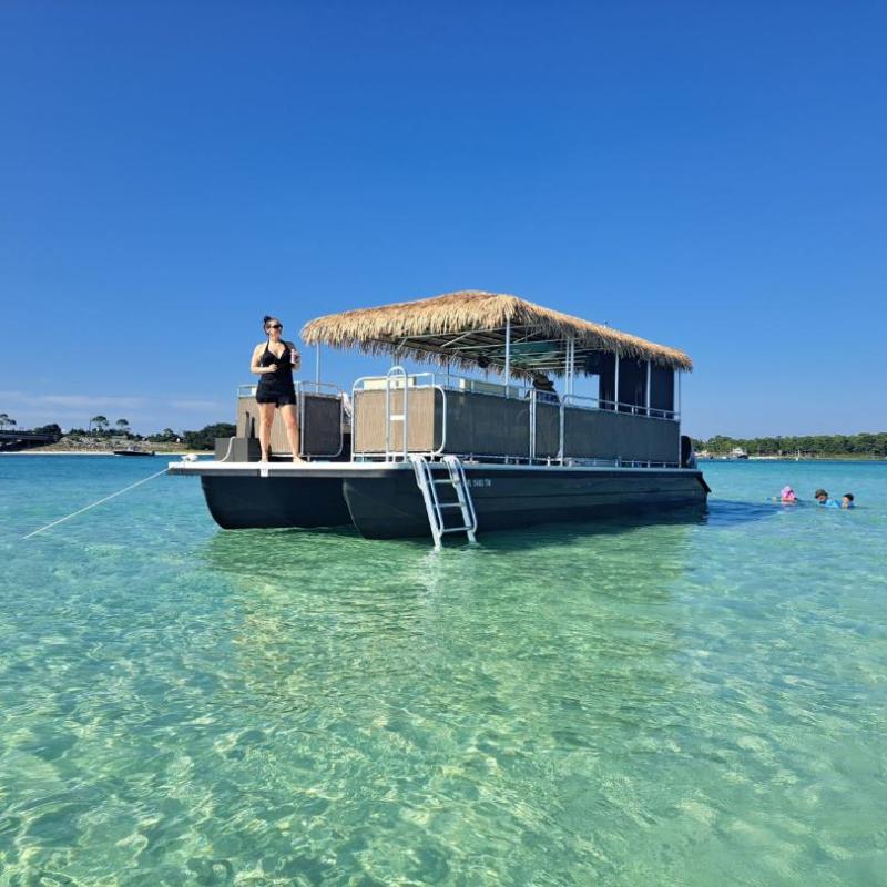 Person on tiki-style pontoon boat in clear water, swimmers nearby, under a clear blue sky.