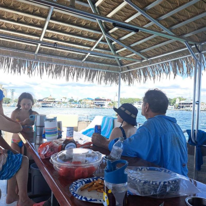 People sitting under a shaded canopied area on a boat with water and buildings in the background.