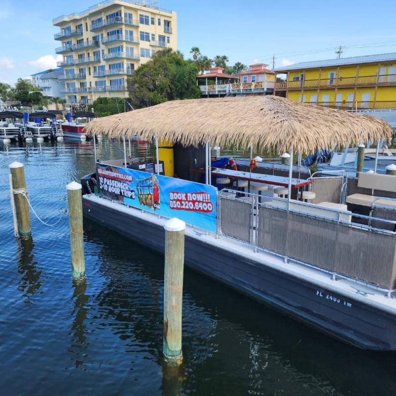 Boat with tiki roof docked near colorful buildings and other boats in a marina.