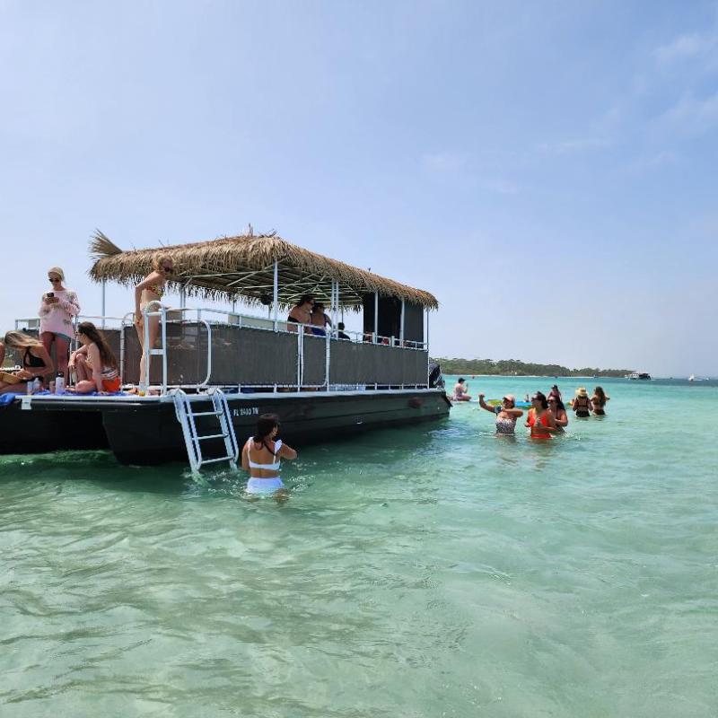 People on a floating barge in shallow, clear ocean water, with others swimming nearby, under a blue sky.