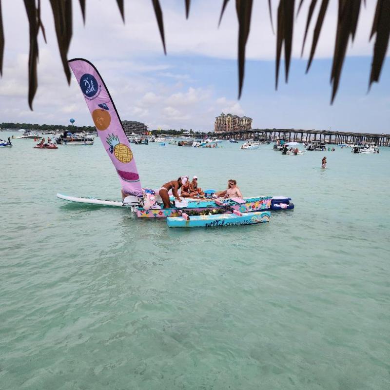 People relaxing on a colorful floating platform in clear water with boats and a bridge in the background.