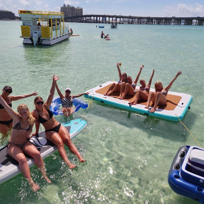People enjoy floating on paddle boards and inflatables in clear water near a boat and bridge.